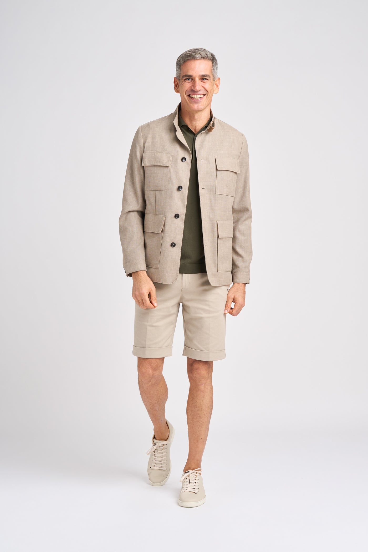 A man wears a beige wool jacket over an olive shirt, paired with light shorts and sneakers.