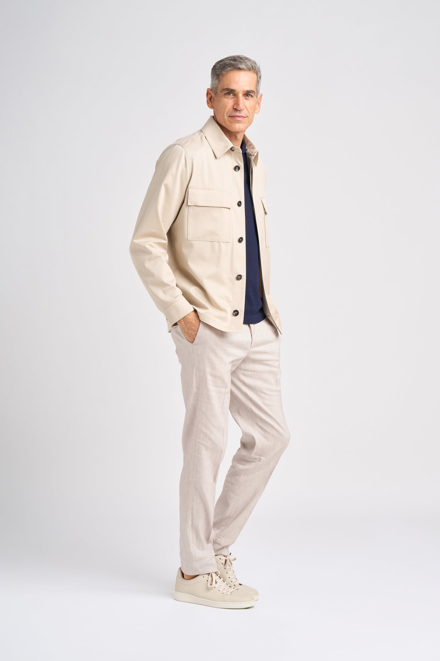 A middle-aged man in a stylish Trofeo cotton and wool Norat jacket and navy top stands confidently with one hand in his pocket, complemented by light linen trousers and beige sneakers against a plain background.