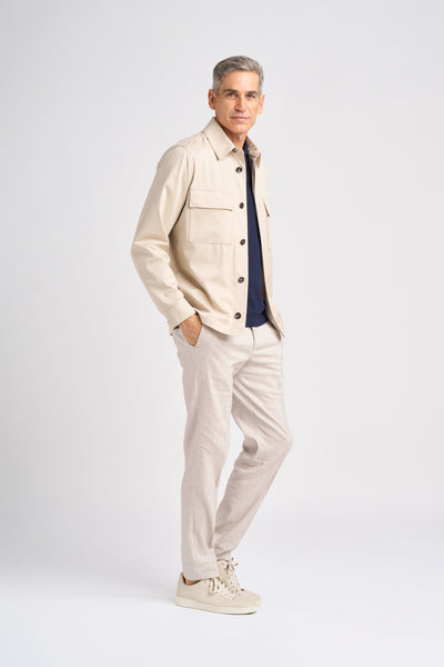 A middle-aged man in a stylish Trofeo cotton and wool Norat jacket and navy top stands confidently with one hand in his pocket, complemented by light linen trousers and beige sneakers against a plain background.