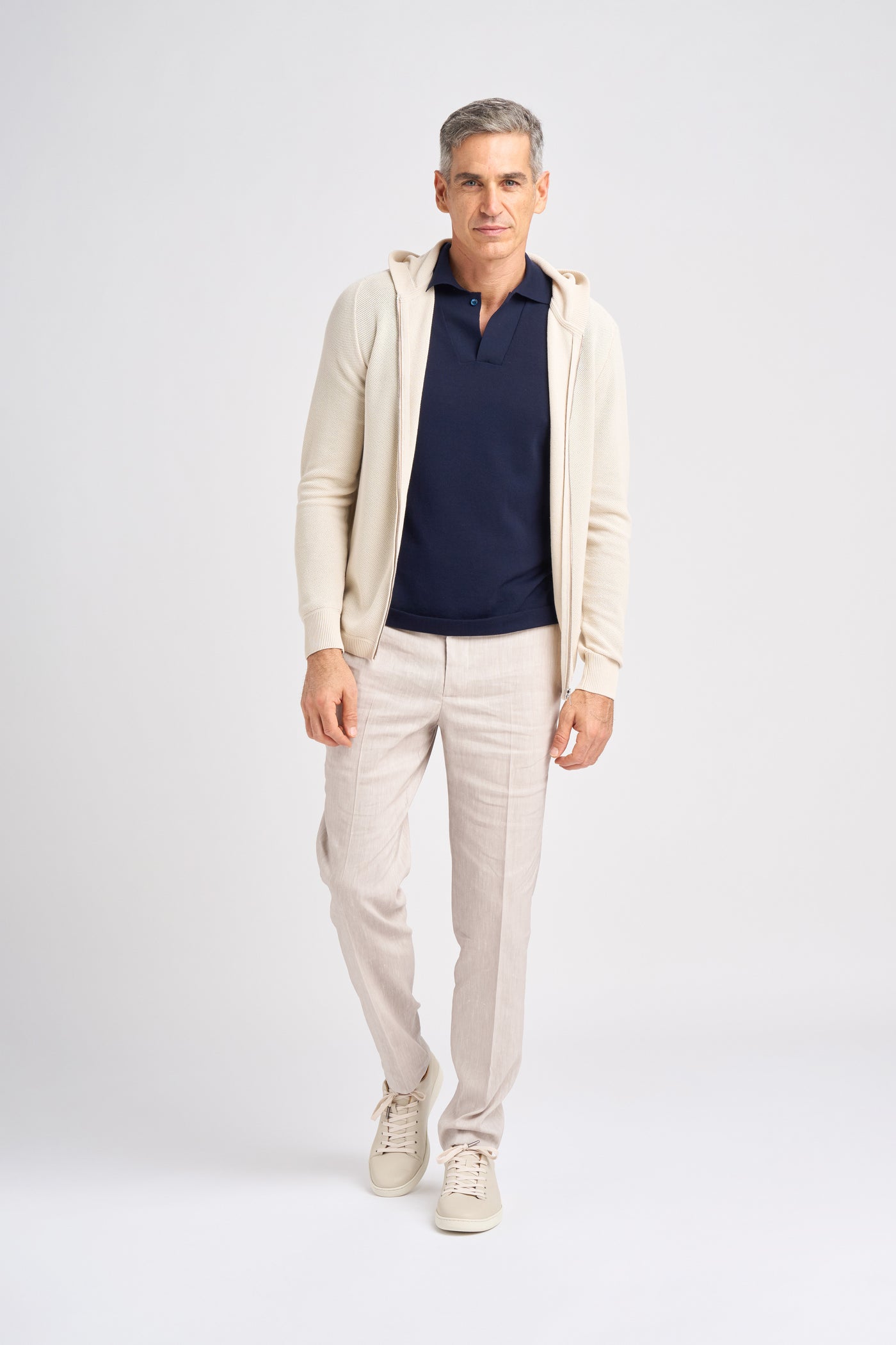 A male model poses in a light-colored Lesien full zip hoodie in Egyptian Cotton over a navy polo shirt, paired with light linen trousers and beige sneakers.