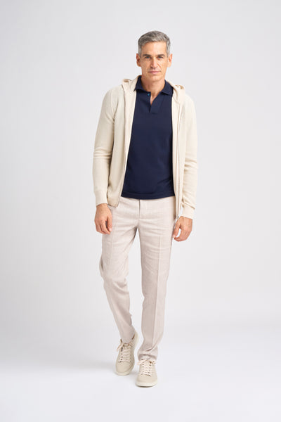 A male model poses in a light-colored Lesien full zip hoodie in Egyptian Cotton over a navy polo shirt, paired with light linen trousers and beige sneakers.