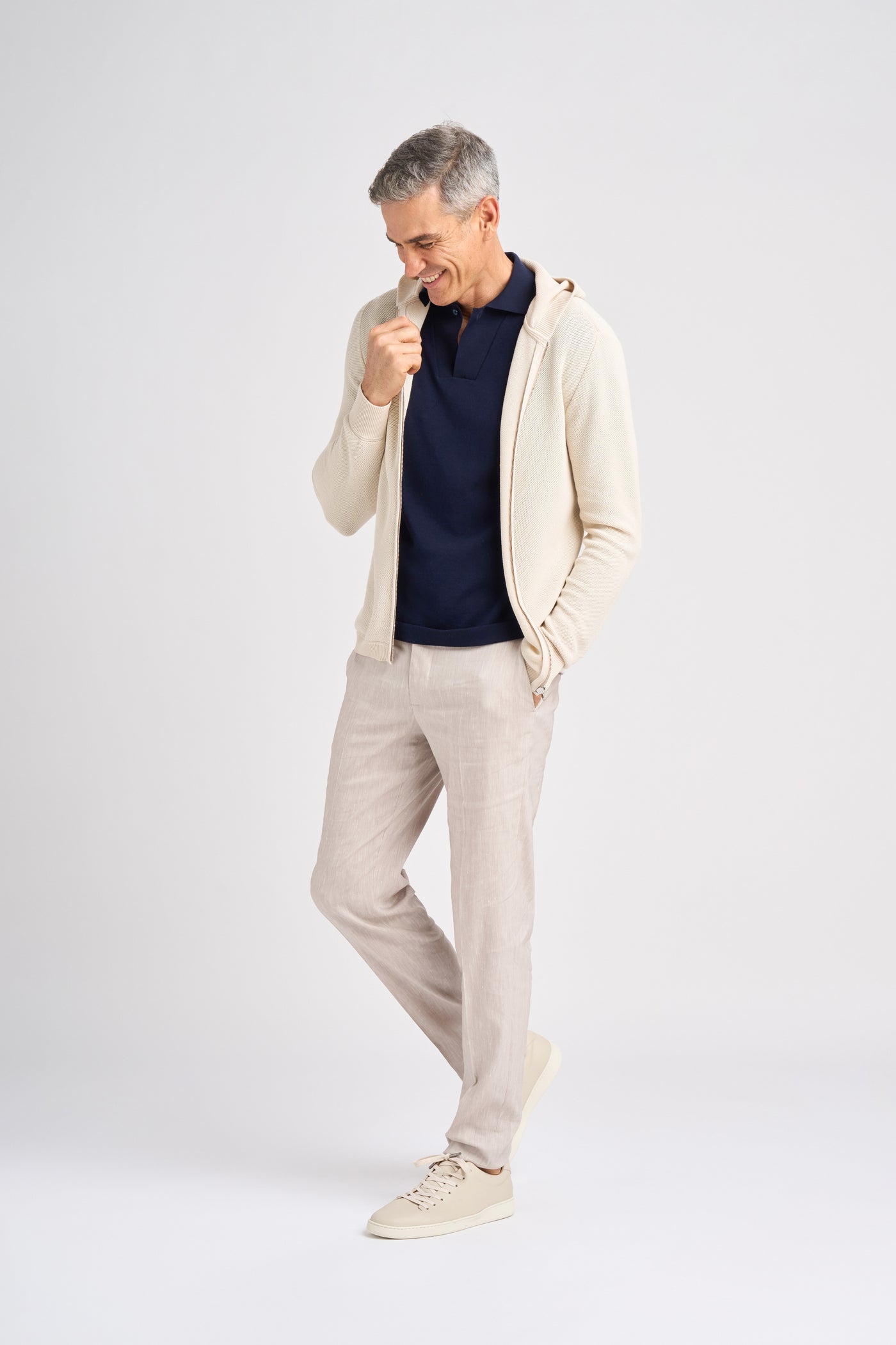 A middle-aged male model smiles while wearing a beige zip-up hoodie over a navy polo shirt, paired with light-colored linen pants and beige sneakers.