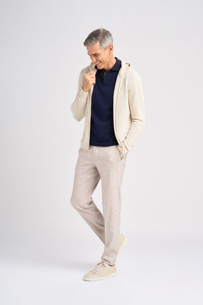 A middle-aged male model smiles while wearing a beige zip-up hoodie over a navy polo shirt, paired with light-colored linen pants and beige sneakers.